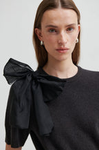 Woman wearing a black sweater with a large bow detail on a plain background