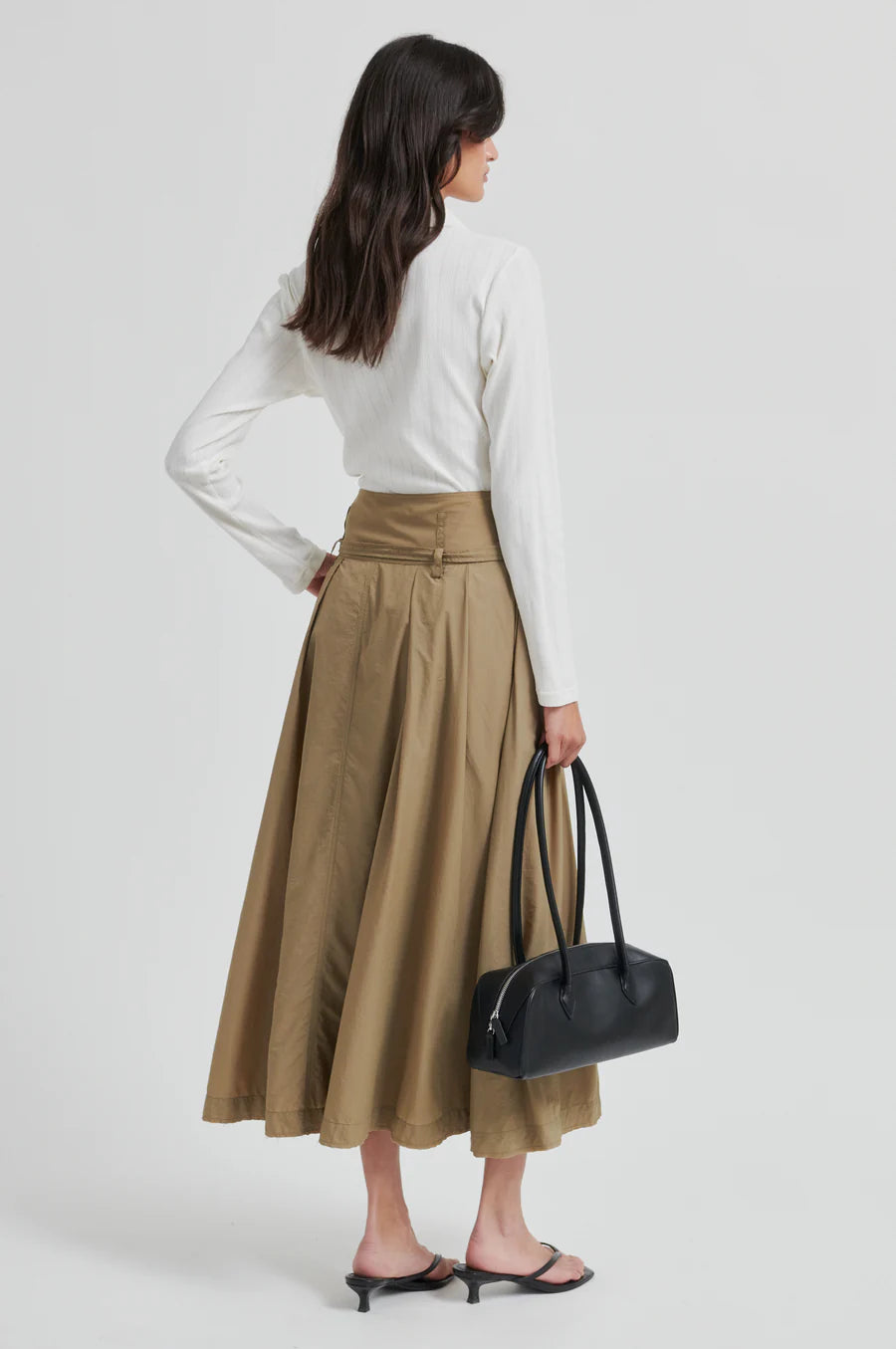 Woman wearing a white blouse and beige skirt, holding a black handbag on a white background