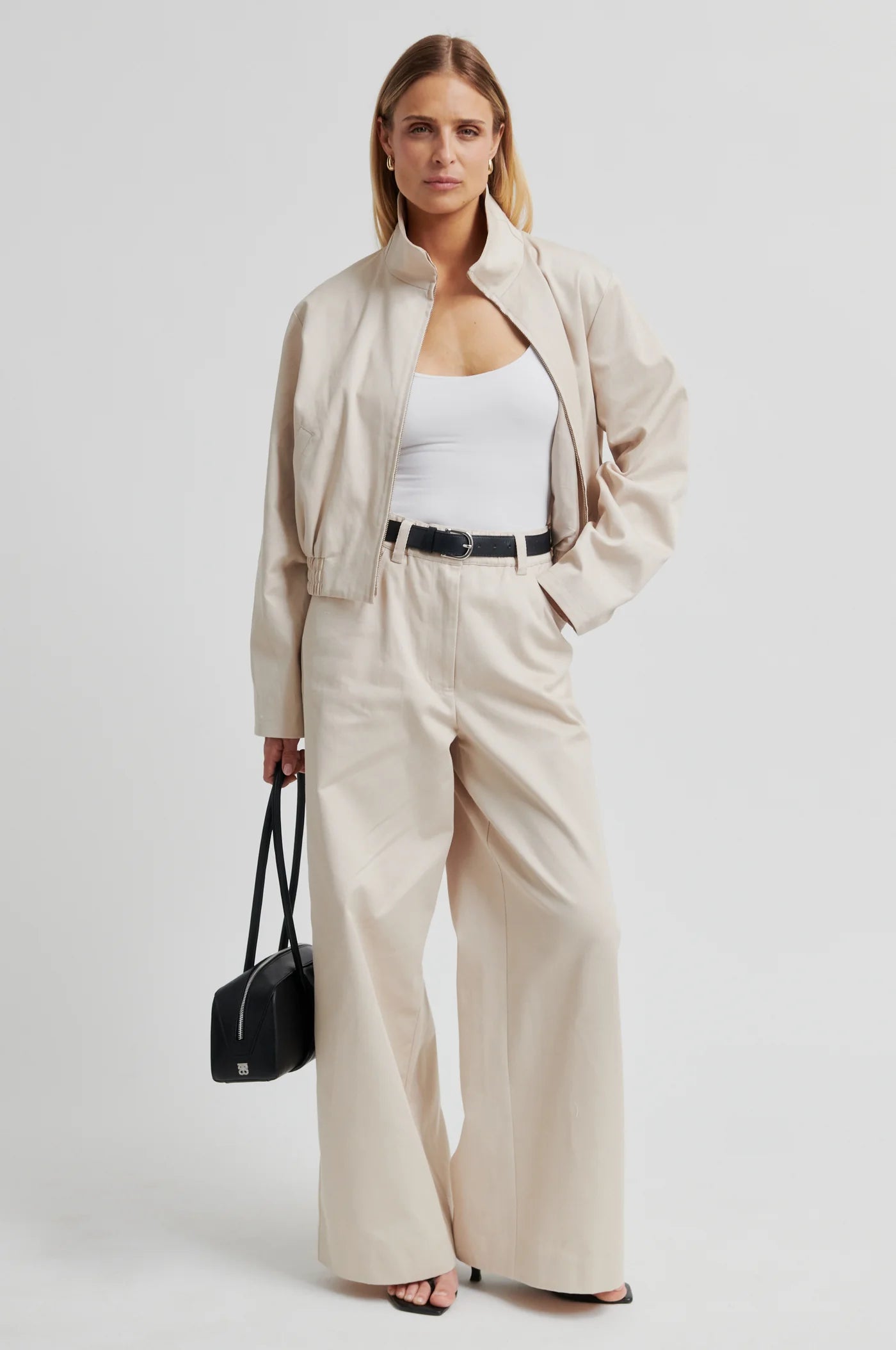Woman wearing a beige outfit with wide-leg pants and a black bag on a white background