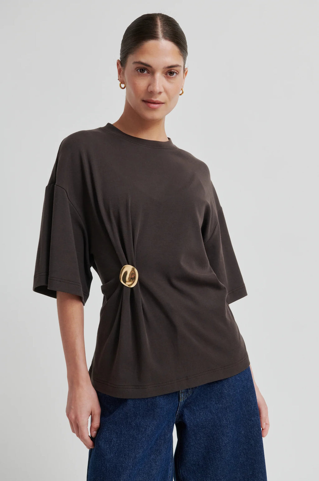 Woman wearing a brown oversized t-shirt with a gold belt and blue jeans on a light gray background