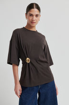 Woman wearing a brown oversized t-shirt with a gold belt and blue jeans on a light gray background