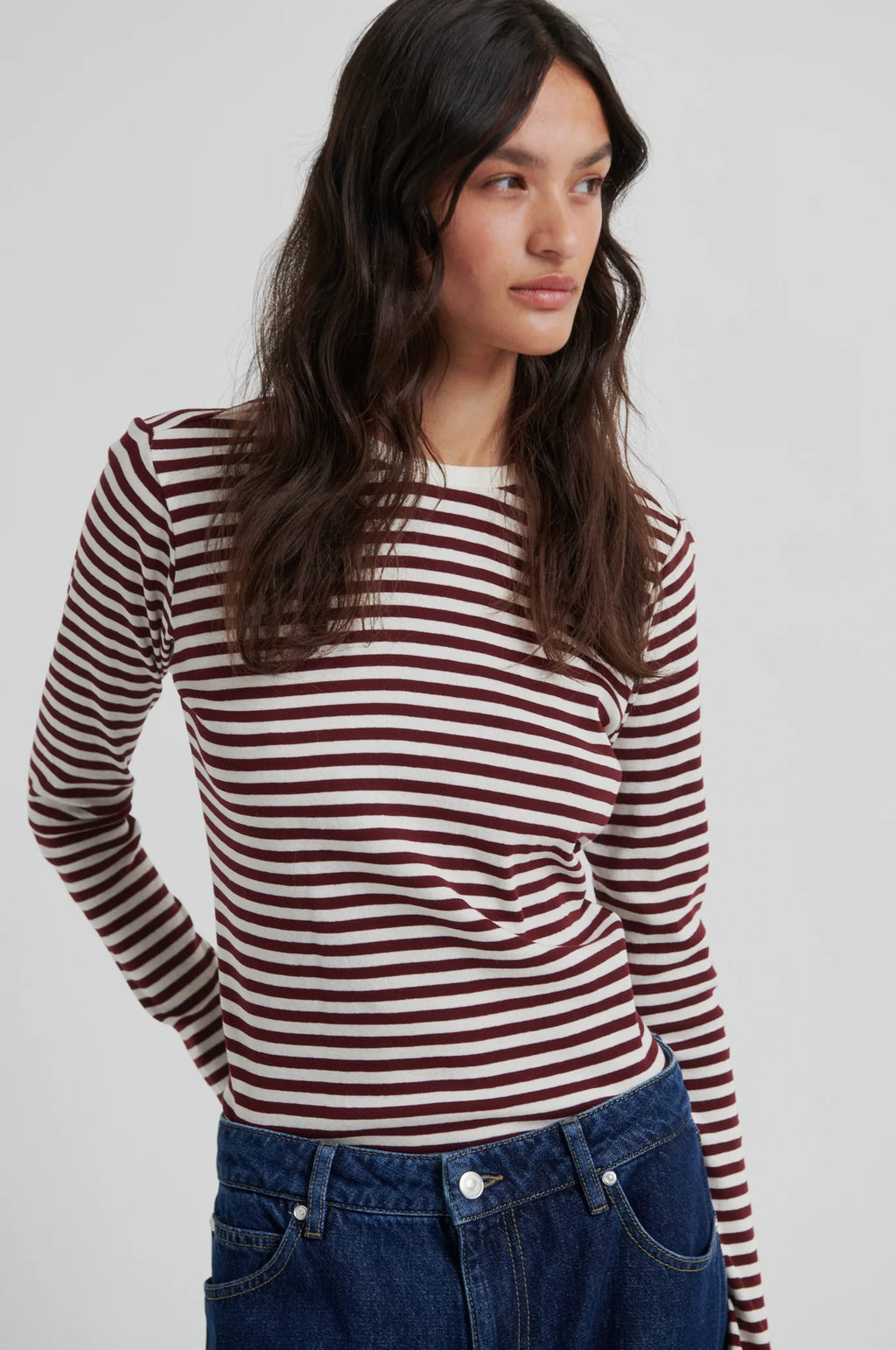 A woman wearing a long-sleeve knit top with horizontal burgundy and off-white stripes, paired with blue jeans, photographed against a light gray studio background.