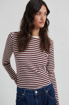 A woman wearing a long-sleeve knit top with horizontal burgundy and off-white stripes, paired with blue jeans, photographed against a light gray studio background.