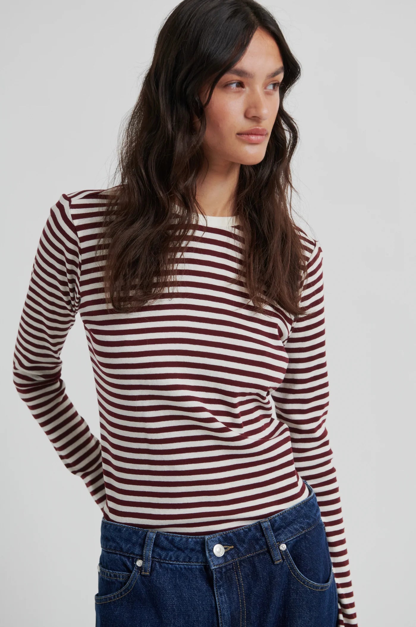 A woman wearing a long-sleeve knit top with horizontal burgundy and off-white stripes, paired with blue jeans, photographed against a light gray studio background.