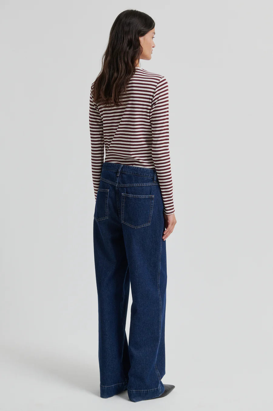 A woman is shown from the back wearing a long-sleeve striped knit top with dark blue wide-leg jeans against a light gray studio background.