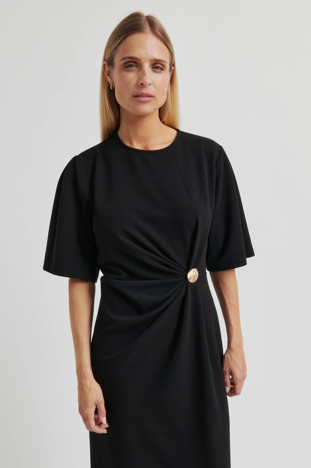 A woman modeling a black short-sleeve midi dress with a twisted waist detail and a single gold button accent, photographed against a light gray studio background.