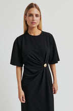 A woman modeling a black short-sleeve midi dress with a twisted waist detail and a single gold button accent, photographed against a light gray studio background.