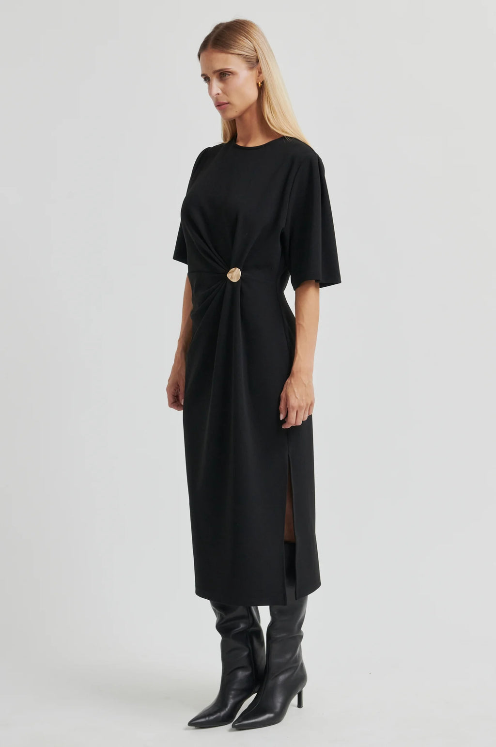 A woman modeling a black midi dress with short sleeves, a gathered twist detail at the waist, and a front slit, paired with black pointed-toe heeled boots against a light gray studio background.