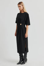 A woman modeling a black midi dress with short sleeves, a gathered twist detail at the waist, and a front slit, paired with black pointed-toe heeled boots against a light gray studio background.