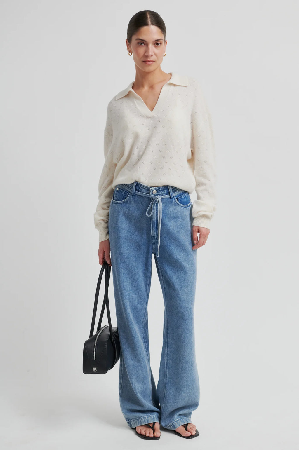 Woman wearing a white sweater and blue jeans holding a black handbag on a white background