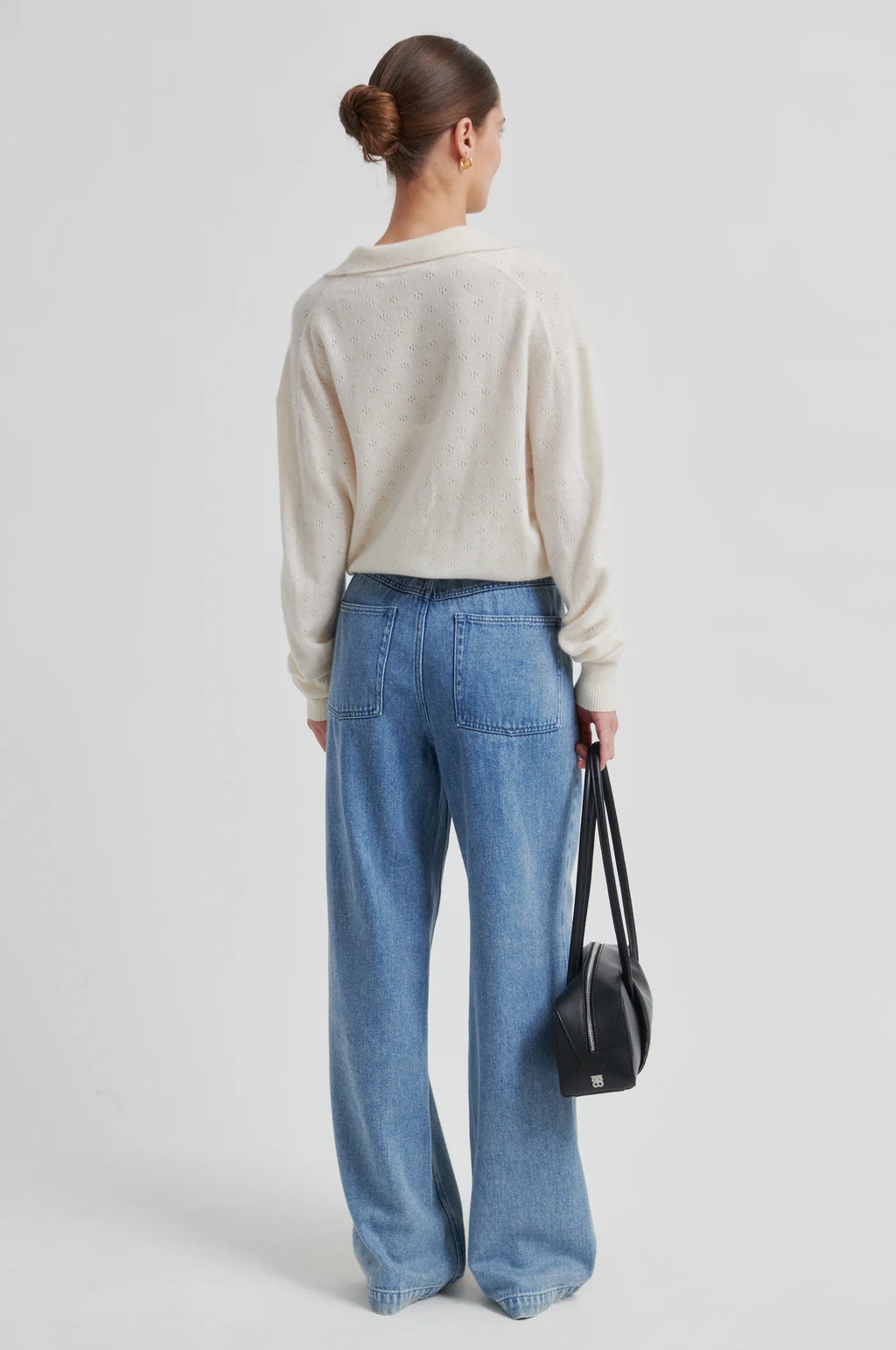 Person wearing a beige sweater and blue jeans holding a black bag on a light gray background