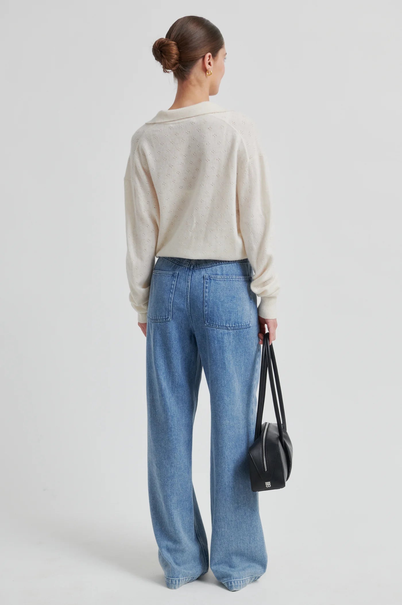 Person wearing a beige sweater and blue jeans holding a black bag on a light gray background