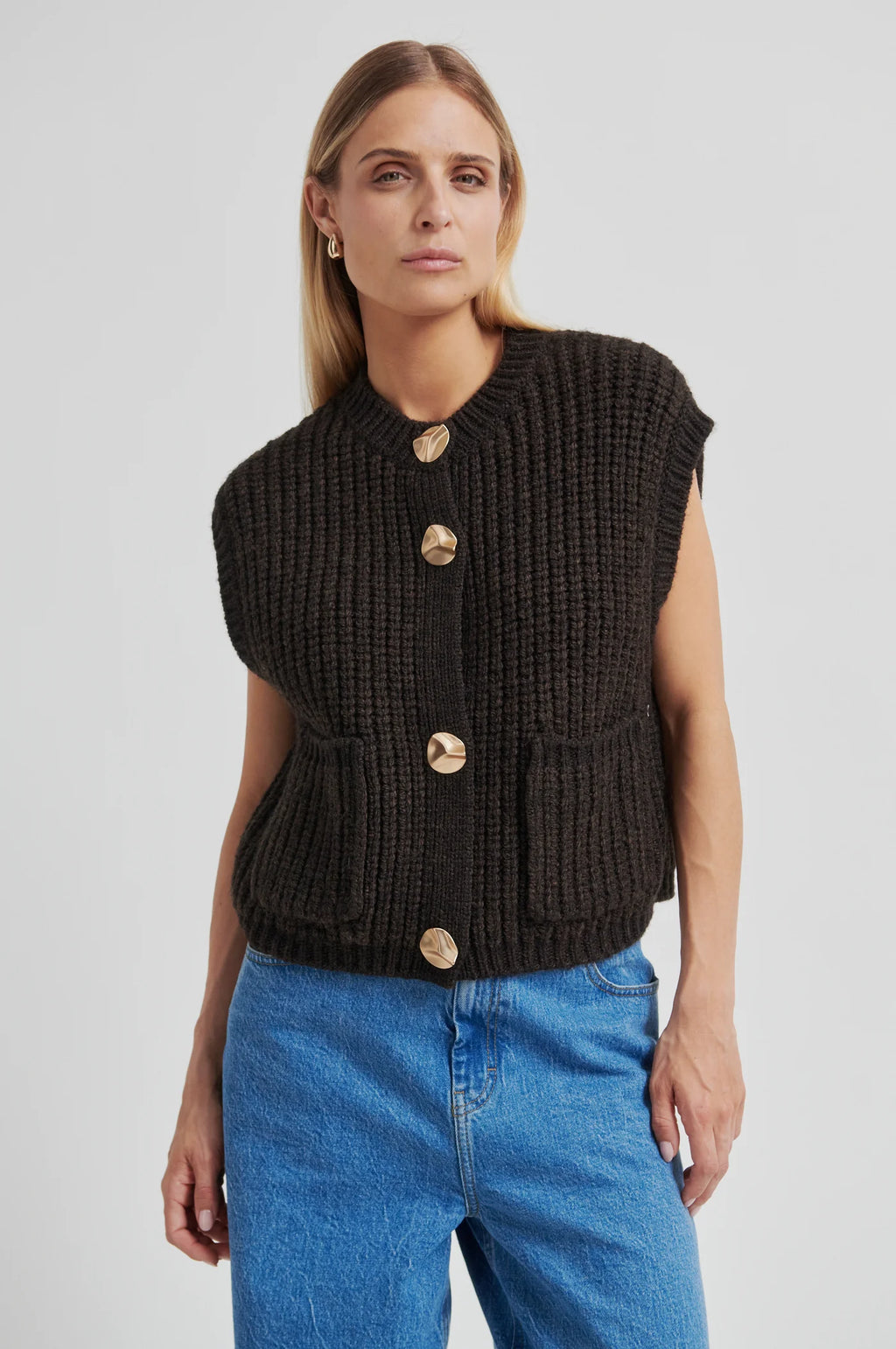 Woman wearing a black knitted vest with gold buttons and blue jeans on a light gray background