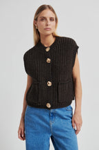 Woman wearing a black knitted vest with gold buttons and blue jeans on a light gray background