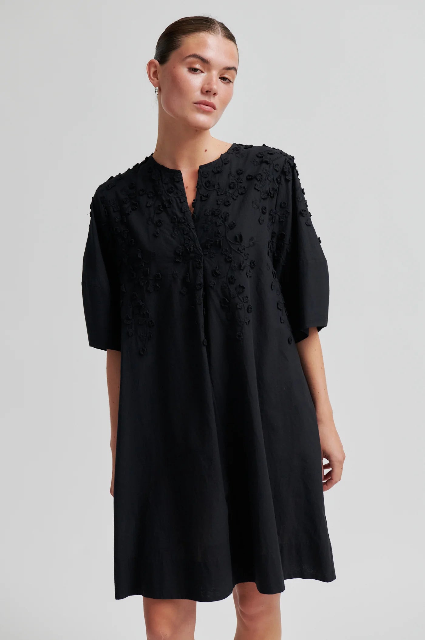 A woman modeling a black short-sleeve A-line dress with floral embroidery on the bodice, photographed against a plain light gray studio background.
