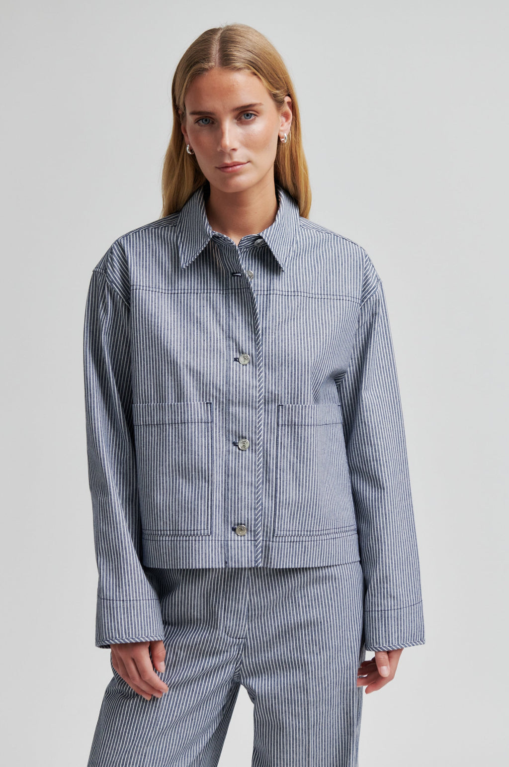 A woman modeling a matching blue and white striped button-up shirt jacket and trousers set against a plain light gray studio background.