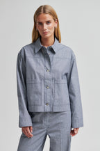 A woman modeling a matching blue and white striped button-up shirt jacket and trousers set against a plain light gray studio background.