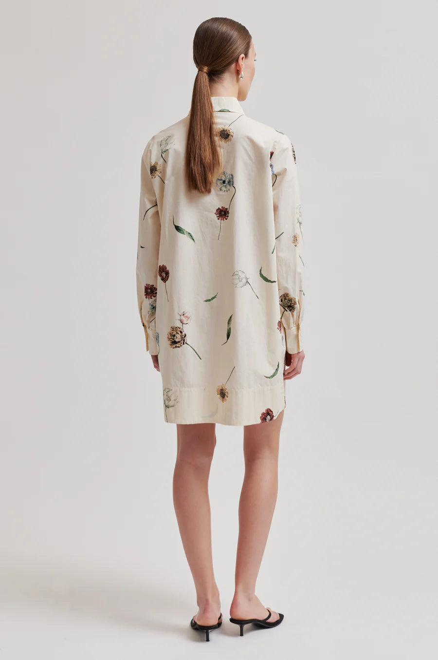 A woman viewed from the back wearing a cream-colored shirt dress with a brown and green floral print, paired with black flat sandals, against a light gray studio background.