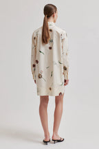A woman viewed from the back wearing a cream-colored shirt dress with a brown and green floral print, paired with black flat sandals, against a light gray studio background.