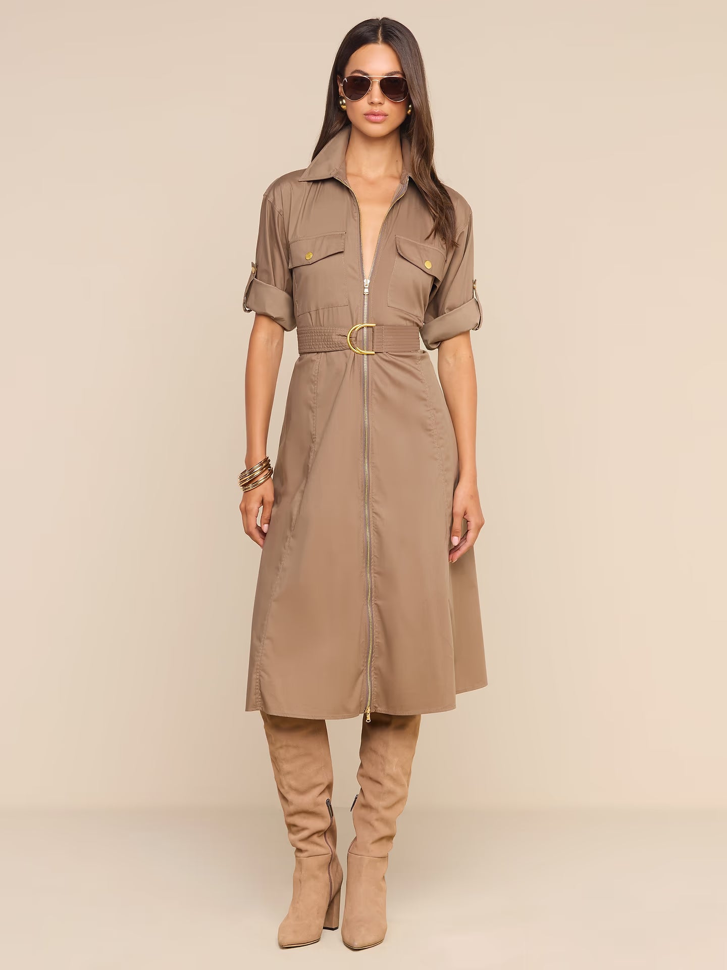 A woman modeling a taupe-brown utility shirt dress with a front zipper, rolled sleeves, flap chest pockets, and a matching belt with a gold buckle, styled with matching boots and sunglasses against a solid beige background.