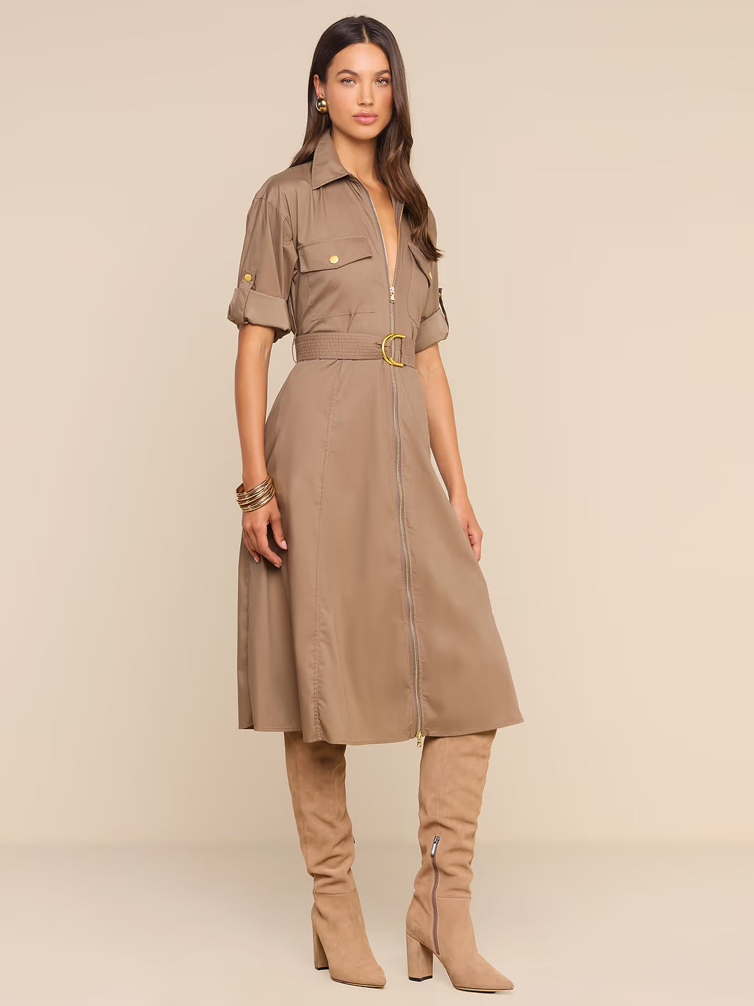 A woman models a taupe-brown midi shirt dress with a front zipper, short sleeves, chest flap pockets, and a matching belt with a gold buckle. She is also wearing matching taupe knee-high boots against a solid beige studio background.