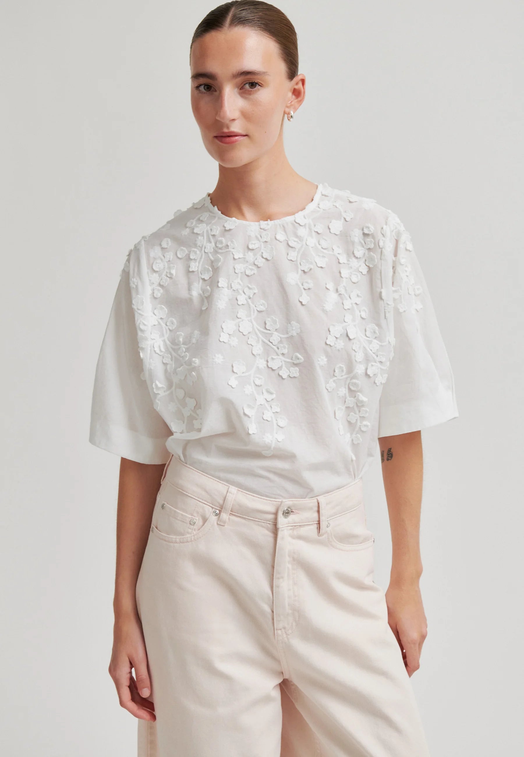 A woman modeling a white short-sleeve blouse with 3D floral embroidery on the bodice, paired with light beige trousers, against a plain light gray studio background.