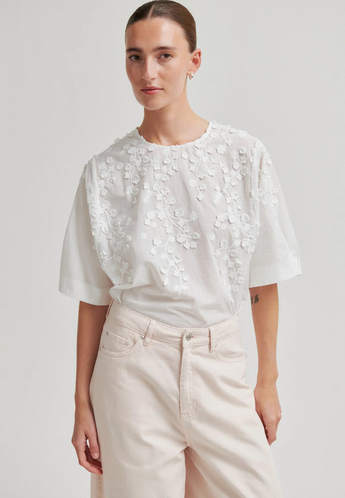 A woman modeling a white short-sleeve blouse with 3D floral embroidery on the bodice, paired with light beige trousers, against a plain light gray studio background.