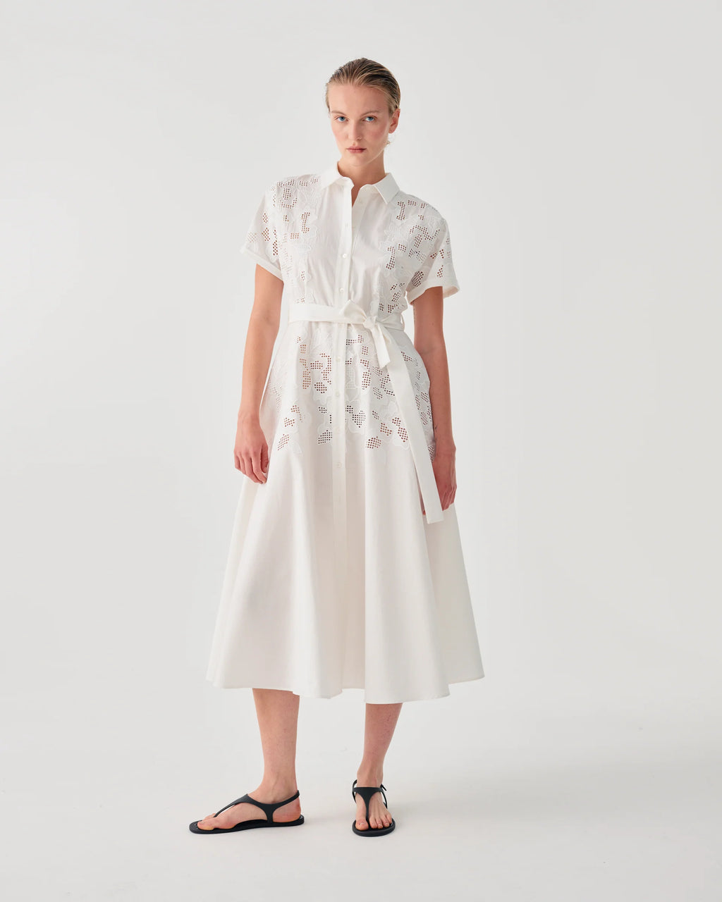 A model wearing a white midi dress with short sleeves, a high collar, and delicate floral embroidery, paired with black flat sandals, set against a plain light gray studio background.