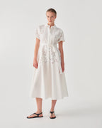 A model wearing a white midi dress with short sleeves, a high collar, and delicate floral embroidery, paired with black flat sandals, set against a plain light gray studio background.