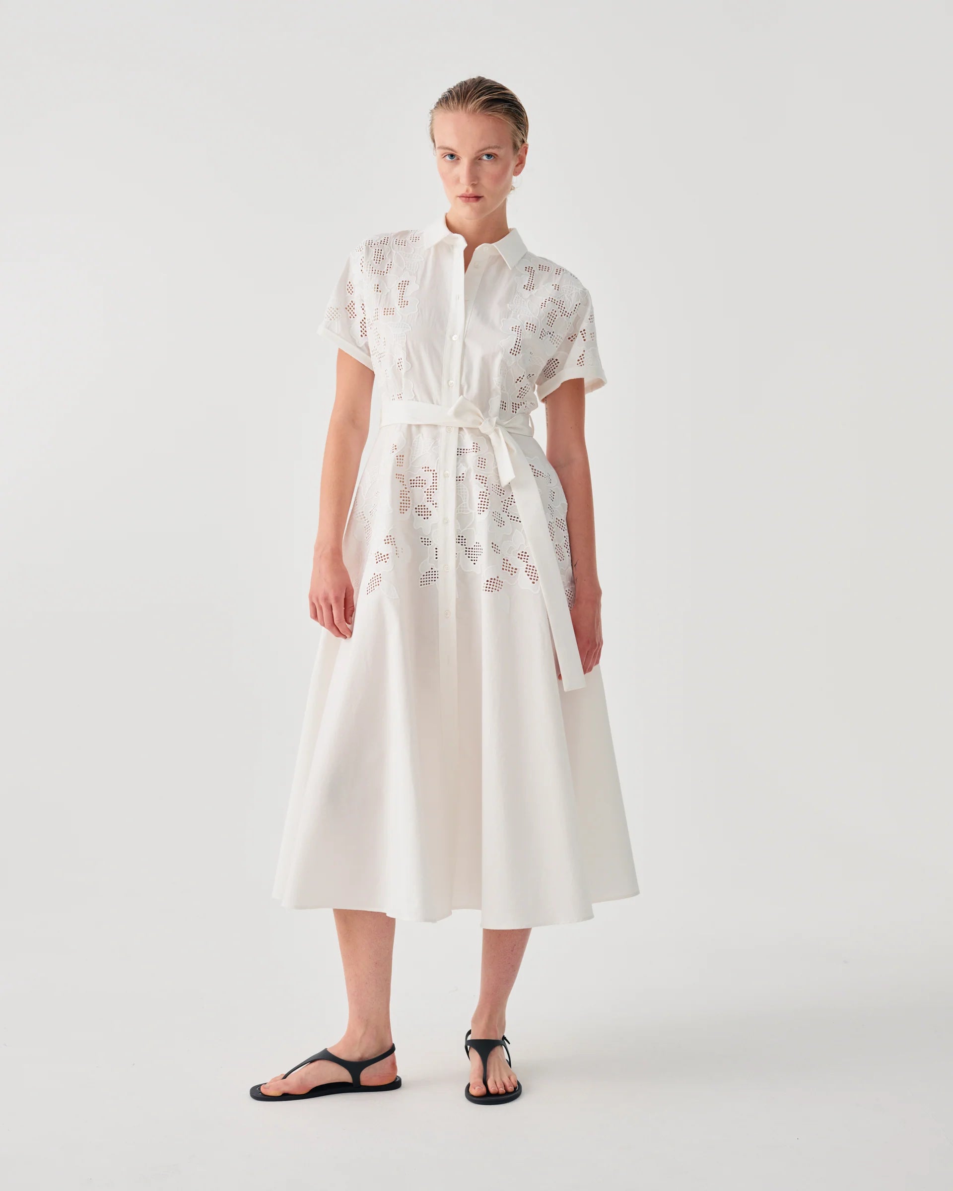 A model wearing a white midi dress with short sleeves, a high collar, and delicate floral embroidery, paired with black flat sandals, set against a plain light gray studio background.