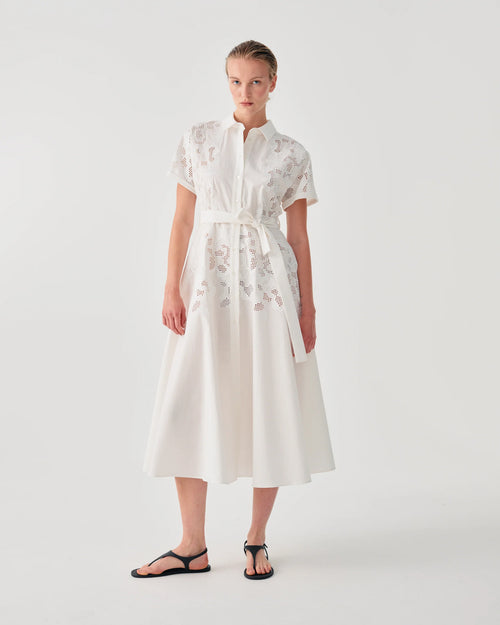 A model wearing a white midi dress with short sleeves, a high collar, and delicate floral embroidery, paired with black flat sandals, set against a plain light gray studio background.