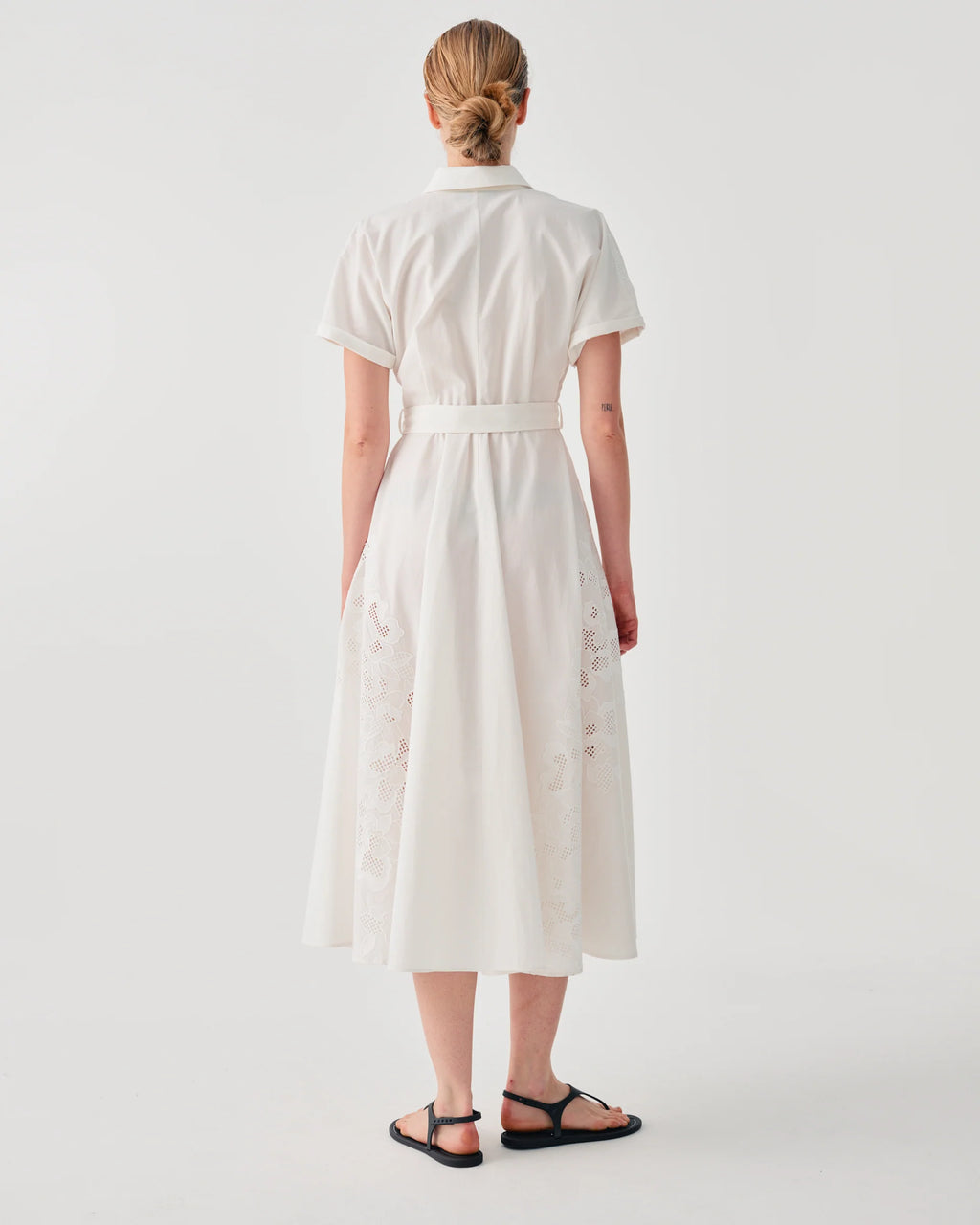 A woman viewed from the back wearing a short-sleeve white midi dress with a matching waist belt and subtle tonal floral embroidery near the hem, paired with black flat sandals against a light gray studio background.