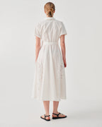 A woman viewed from the back wearing a short-sleeve white midi dress with a matching waist belt and subtle tonal floral embroidery near the hem, paired with black flat sandals against a light gray studio background.