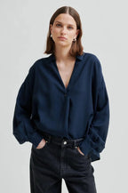 A woman wearing a dark navy blue button-down shirt with a V-neck and long sleeves, paired with dark jeans, photographed against a light gray studio background.
