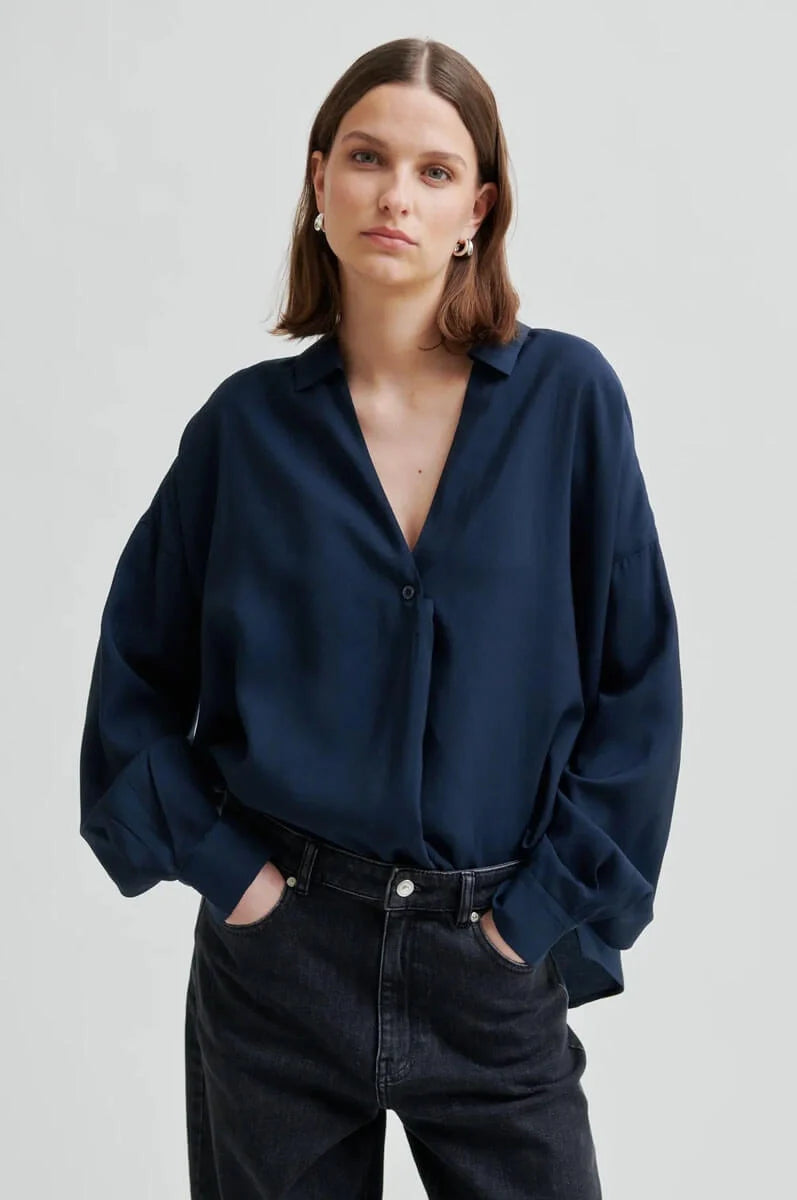 A woman wearing a dark navy blue button-down shirt with a V-neck and long sleeves, paired with dark jeans, photographed against a light gray studio background.