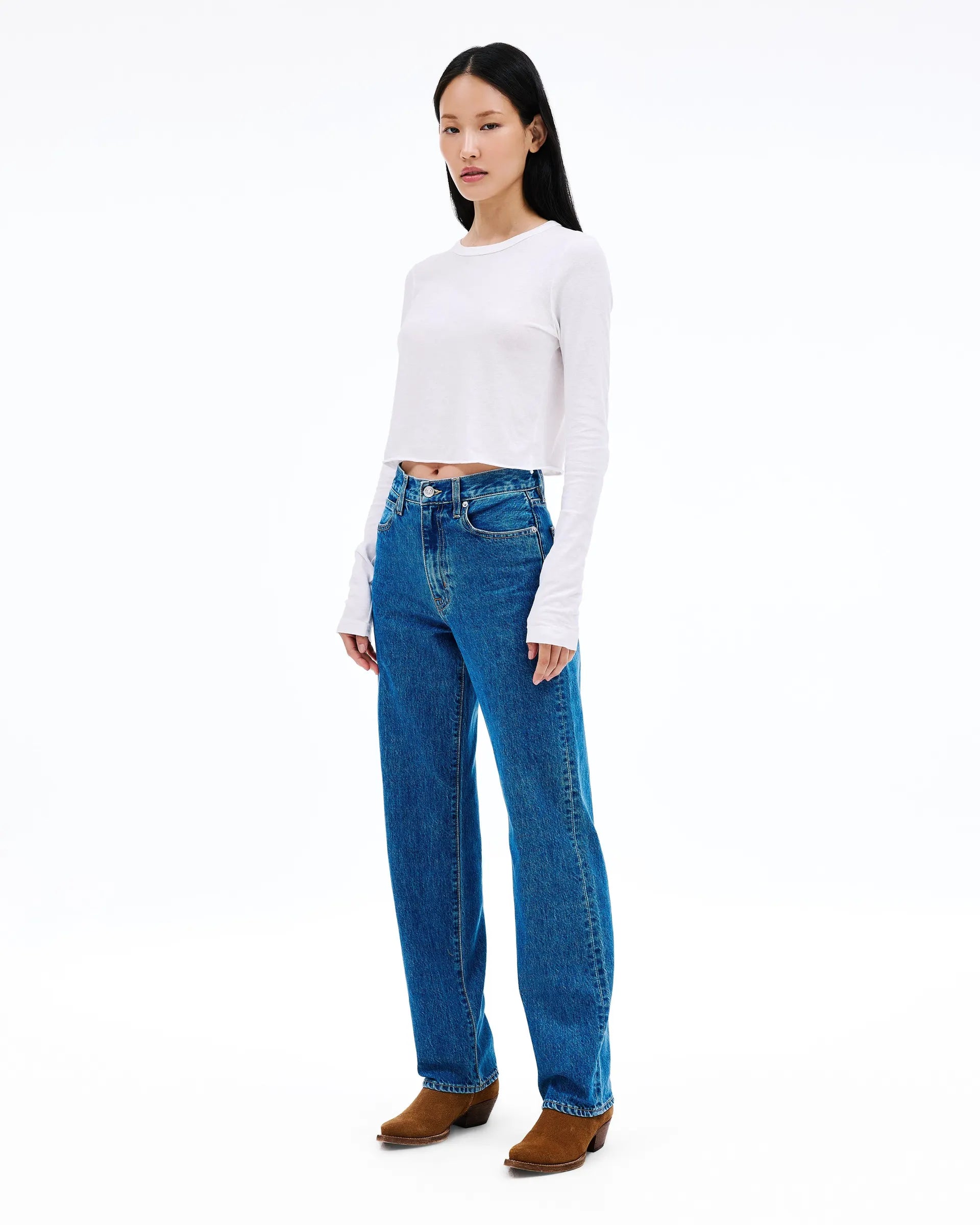 A woman modeling a white long-sleeve cropped top and blue straight-leg jeans with brown boots, standing against a plain white studio background.