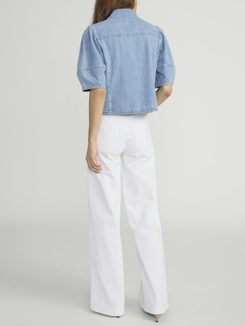 Person wearing a light blue denim shirt and white pants on a plain background