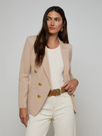 Woman wearing a beige blazer with gold buttons, white top, and white pants on a plain background