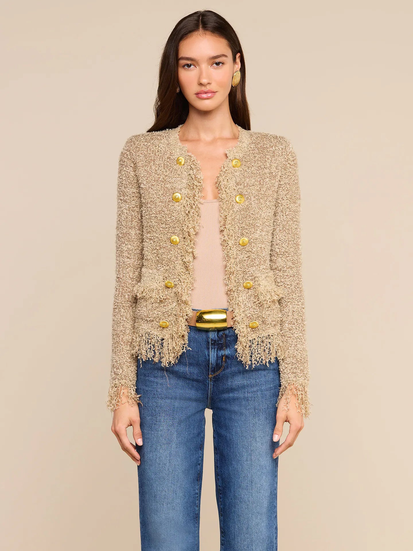 A woman modeling a beige tweed jacket with gold buttons and fringe trim, worn over a light pink top and blue jeans, against a solid beige background.