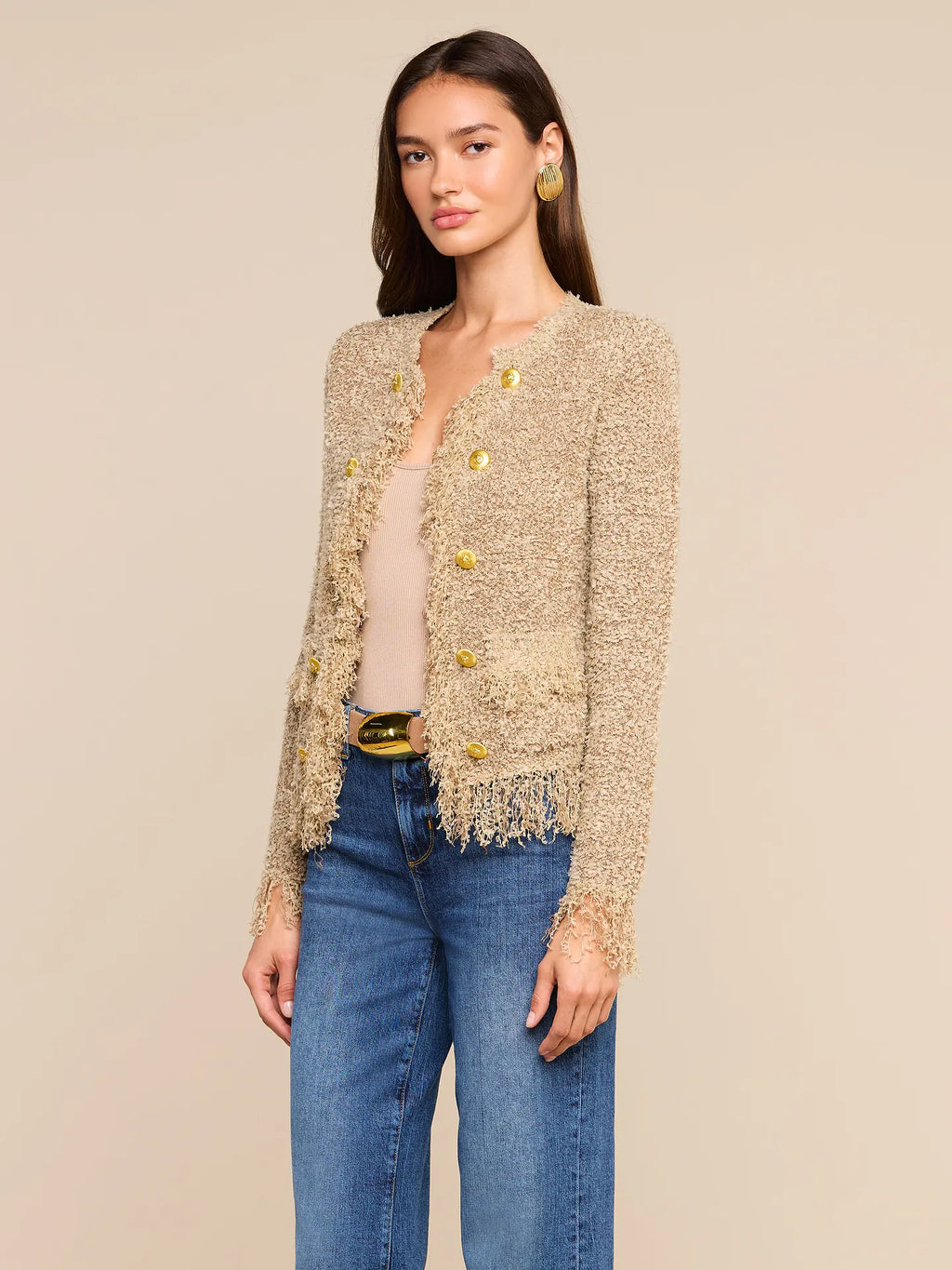 A woman modeling a beige textured tweed cardigan with gold buttons and fringe trim, worn over a beige top and blue jeans, against a solid beige background.
