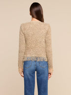 A woman shown from the back wearing a textured beige knit sweater with fringe trim and blue jeans, set against a solid light beige background.