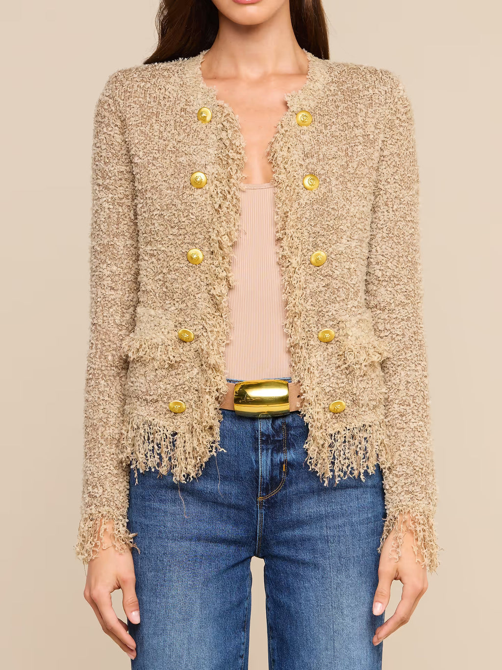 A woman wearing a beige textured fringed jacket with gold buttons, a light pink top, and blue jeans with a gold belt, against a solid beige background.
