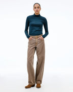 A model stands against a white studio background wearing a dark teal mock-neck long-sleeve top, high-waisted brown wide-leg corduroy pants, and brown suede boots.