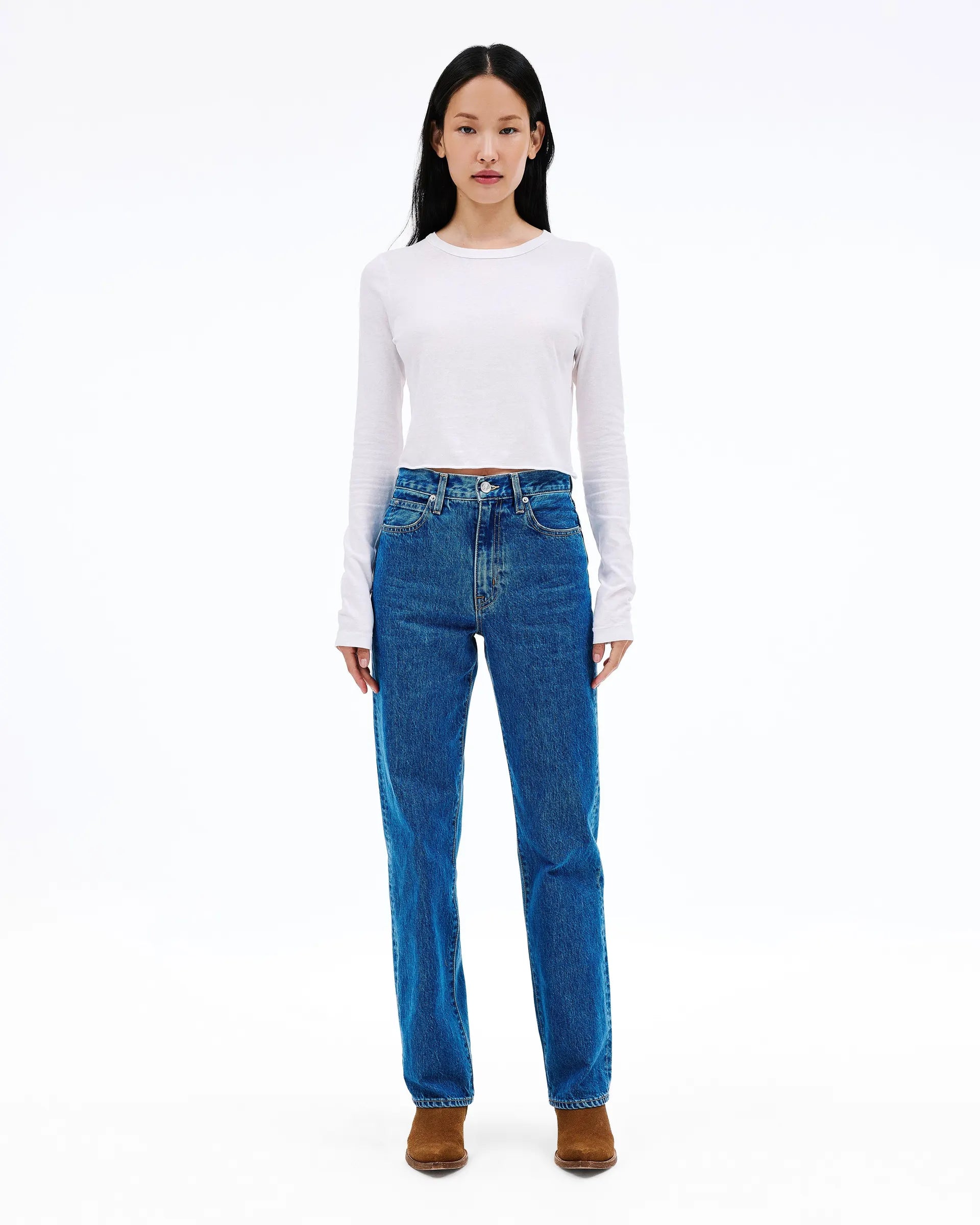 A model stands against a plain white studio background wearing a white long-sleeve cropped top, blue straight-leg jeans, and brown ankle boots.