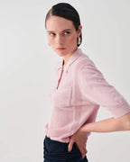 A model wearing a light pink ribbed knit polo shirt with a half-zip collar and elbow-length sleeves, paired with dark trousers, against a plain light gray studio background.