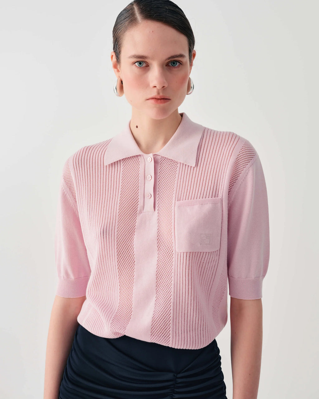 A model wearing a light pink short-sleeve knit polo shirt with a textured vertical stripe pattern and a chest pocket, paired with a dark skirt against a light gray background.