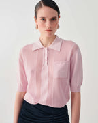 A model wearing a light pink short-sleeve knit polo shirt with a textured vertical stripe pattern and a chest pocket, paired with a dark skirt against a light gray background.
