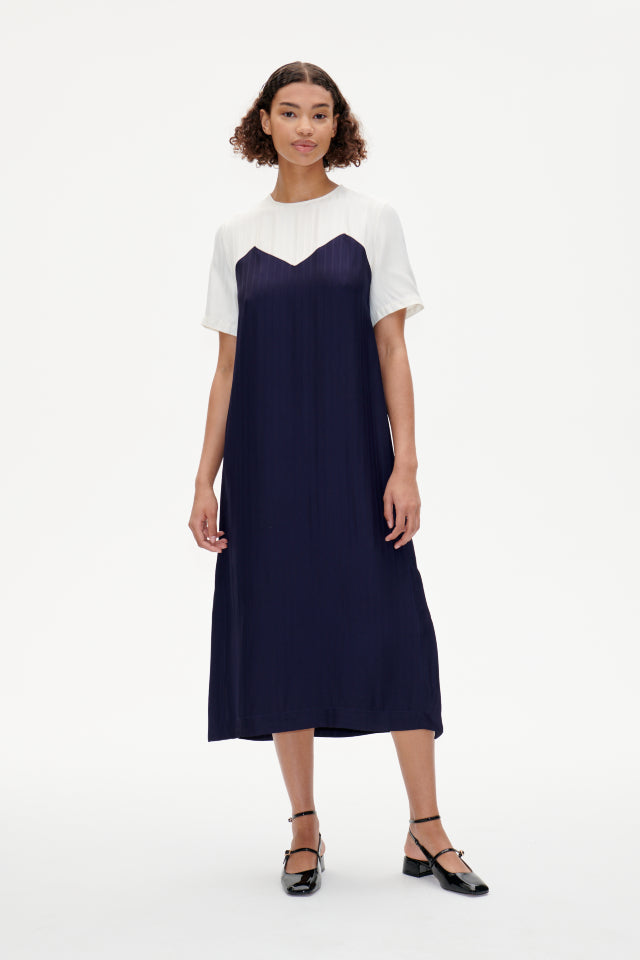 A model wearing a navy blue, midi-length dress with button closures at the neck and slits on the sides. The dress features a sweetheart-style pattern at the chest.