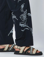 Person wearing black pants with white floral embroidery and sandals on a light gray background