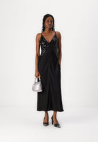 A woman standing wearing a black maxi slip dress with a deep V-neck and sequins at the chest, featuring a side zip and pockets. She is also holding a metallic bag and wearing black heels.